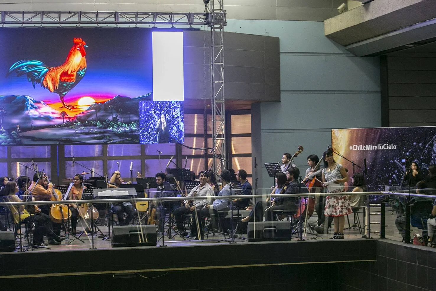 Orquesta de cuerdas toca en un escenario con pantallas LED que muestran un gallo colorido y el hashtag #ChileMiraTuCielo.