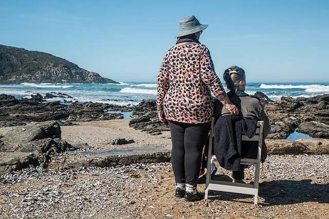 Dos mujeres mayores de espaldas contemplan el mar desde una playa rocosa en un día soleado.