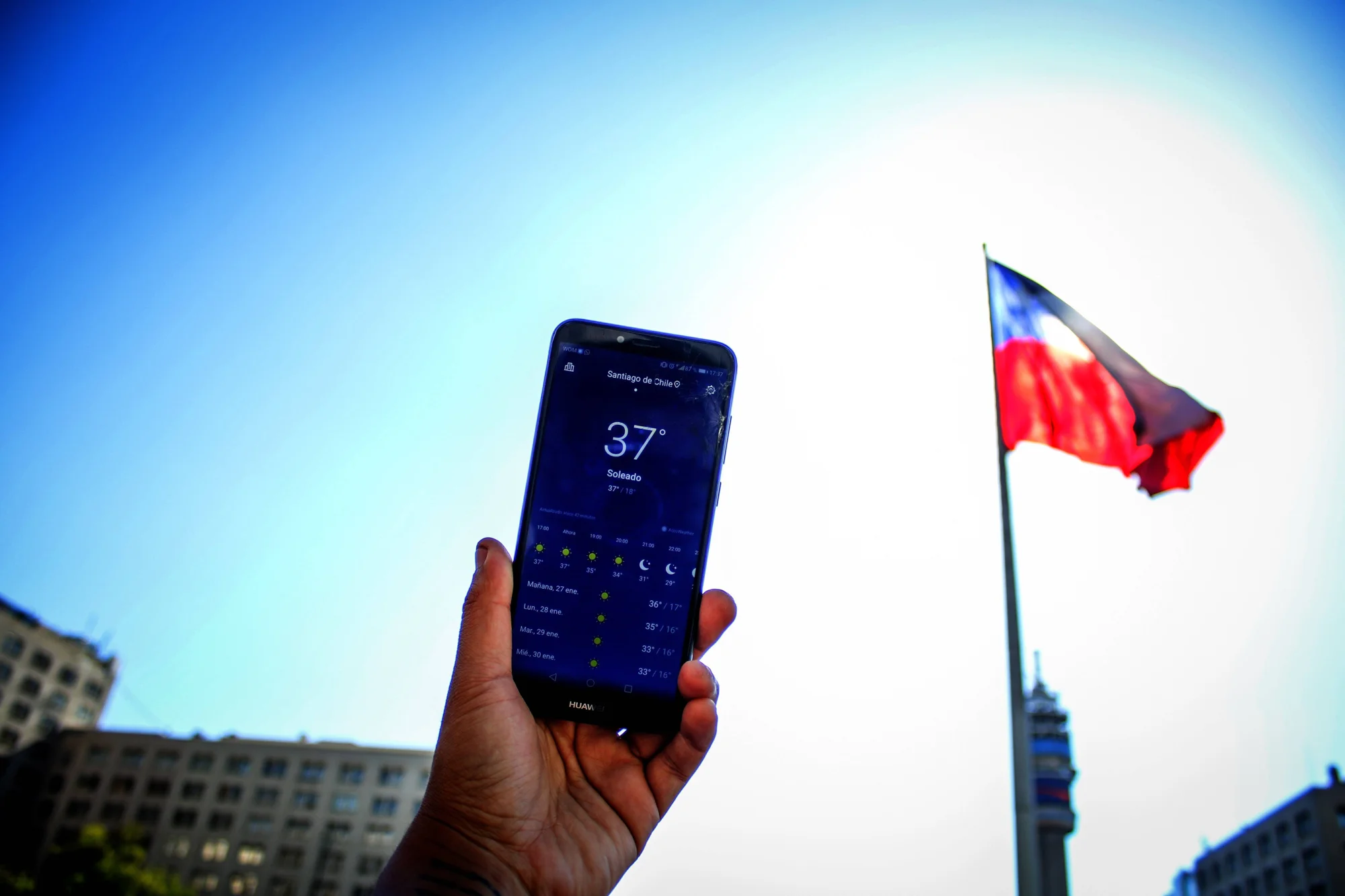 Mano sostiene un smartphone con app del clima mostrando 37°C soleado en Santiago de Chile, con la bandera chilena al fondo.
