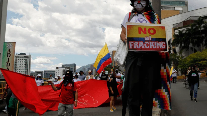Manifestante con vestimenta indígena sostiene un cartel 'No al Fracking' durante una protesta en Colombia, con banderas colombianas y pancartas rojas al fondo.