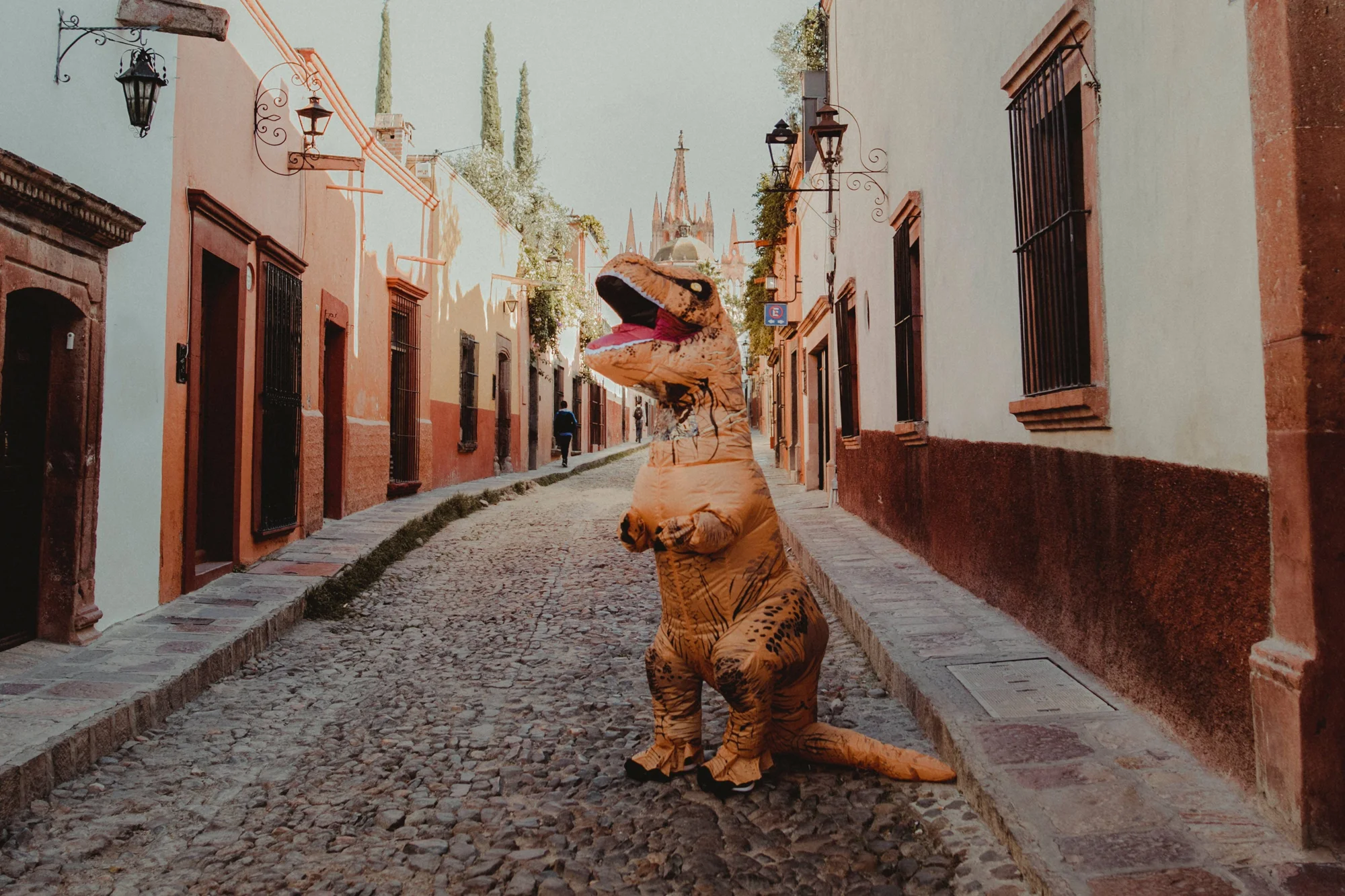 Persona con disfraz inflable de T-Rex parada en una calle empedrada colonial con una iglesia de fondo.
