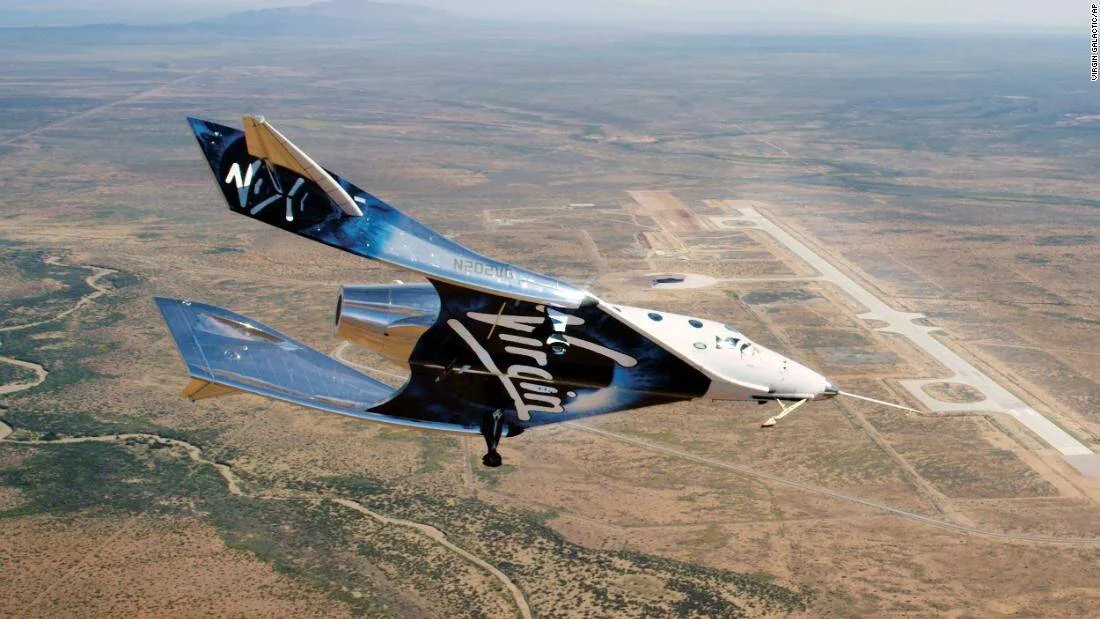 La nave VSS Unity de Virgin Galactic vuela sobre un paisaje desértico con una pista de aterrizaje visible al fondo.
