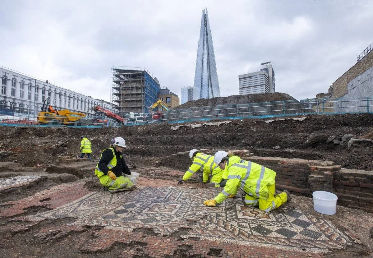 Arqueólogos con chalecos amarillos excavan y limpian un mosaico romano descubierto en una obra urbana de Londres, con el rascacielos The Shard al fondo.