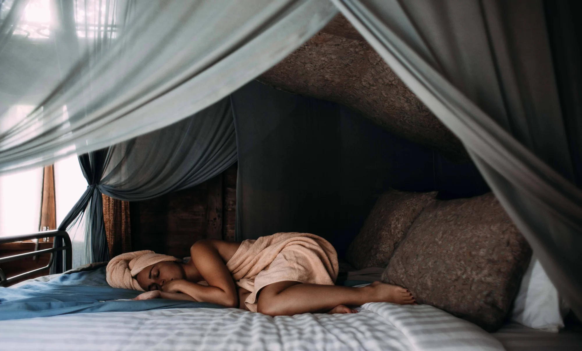 Mujer envuelta en toalla descansa dormida sobre una cama con dosel de cortinas grises en habitación oscura.