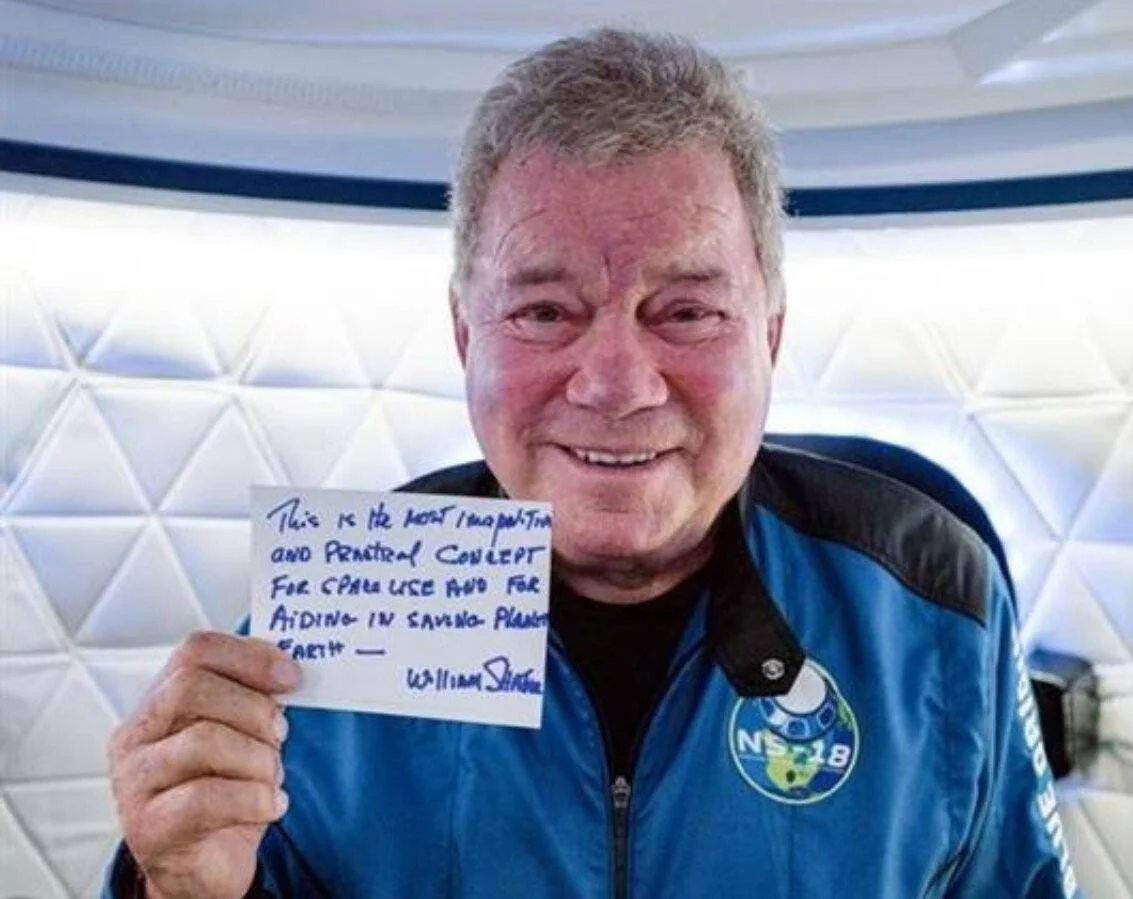 Hombre mayor con chaqueta de vuelo azul NS-18 sonríe mientras sostiene una nota manuscrita firmada por William Shatner sobre salvar el planeta Tierra.