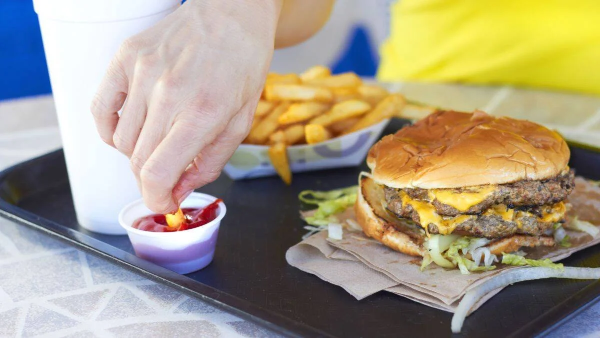 Mano mojando una papa frita en kétchup junto a una hamburguesa doble con queso y papas fritas en una bandeja.