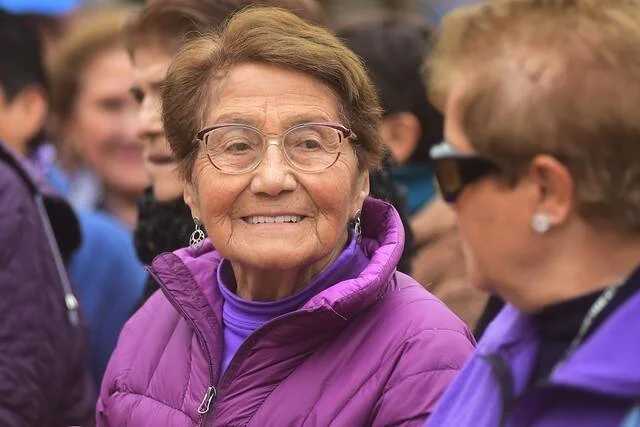 Mujer adulta mayor con anteojos y campera violeta sonríe entre otras personas en un evento al aire libre.