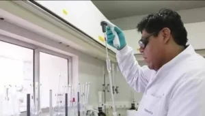 Investigador con bata blanca y guantes aplica una micropipeta sobre material de vidrio en un laboratorio.