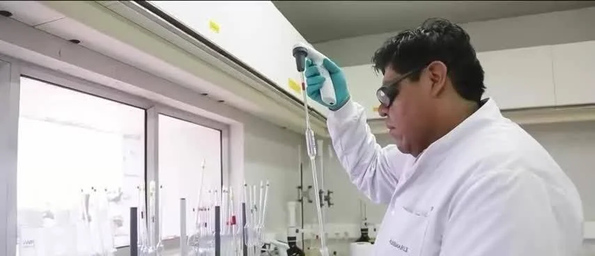 Investigador con bata blanca y guantes aplica una micropipeta sobre material de vidrio en un laboratorio.