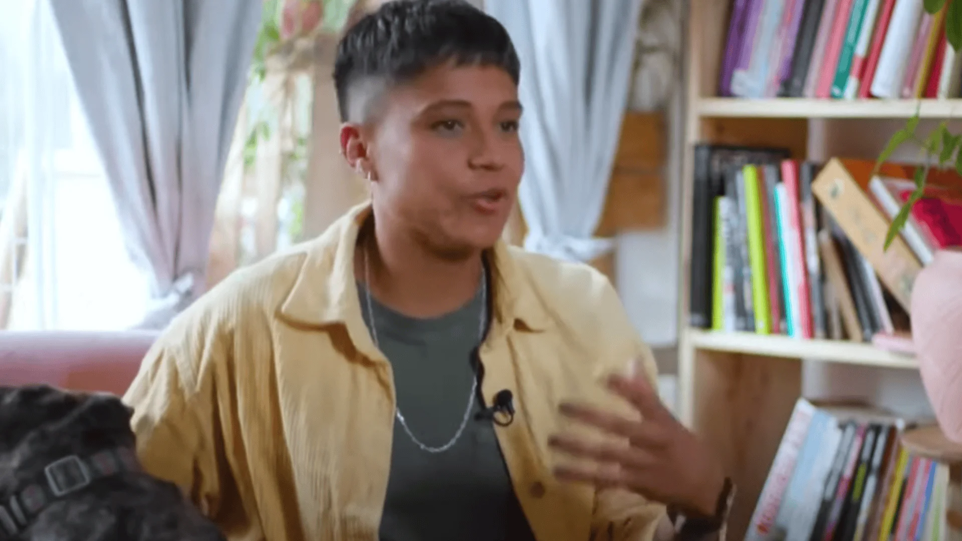 Persona joven con camisa amarilla y micrófono de solapa gesticula mientras habla frente a una estantería con libros.