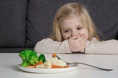 Niña pequeña cubre su boca con las manos frente a un plato de verduras que no quiere comer.