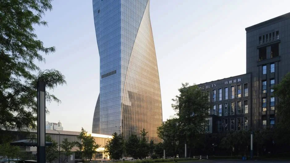 Rascacielos de fachada de vidrio con forma torsionada se eleva entre árboles y edificios de menor altura en una ciudad china.