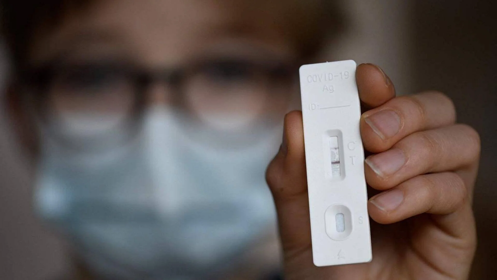 Una persona con mascarilla sostiene un test de antígenos de COVID-19 con resultado positivo visible en dos líneas.