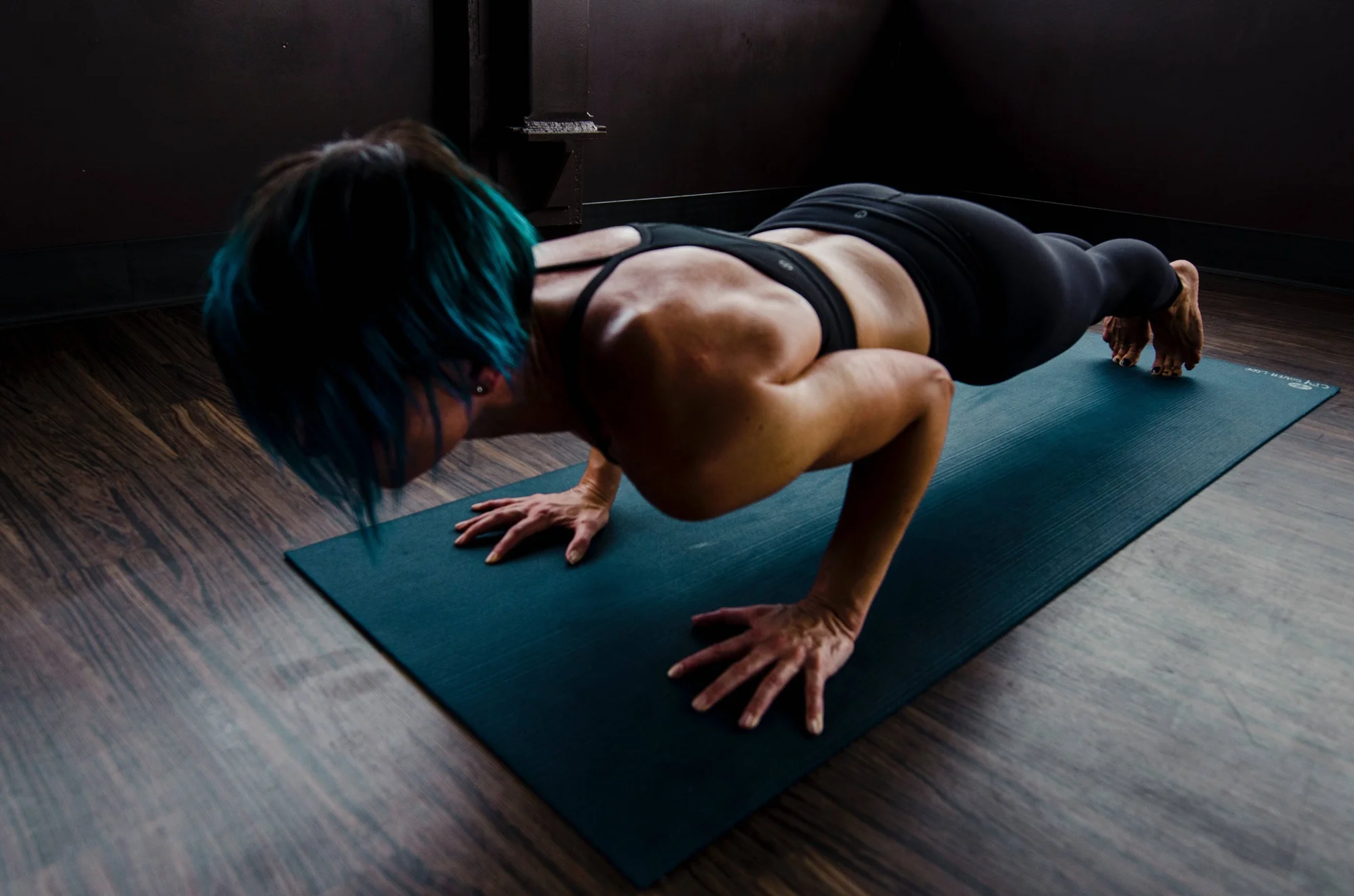 Mujer con cabello azul realiza flexiones de brazos sobre una colchoneta de ejercicio en un gimnasio oscuro.
