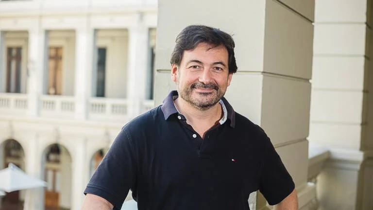 Hombre de mediana edad con remera polo oscura sonríe frente a un edificio de arquitectura clásica con columnas blancas.
