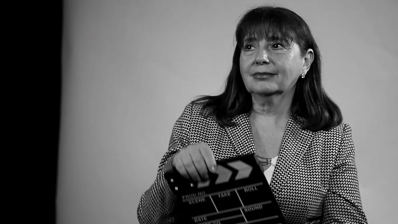 Mujer de cabello oscuro con saco sostiene una claqueta de filmación en una toma en blanco y negro.