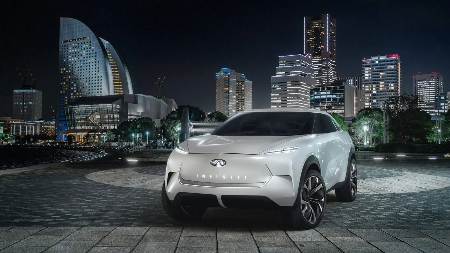 SUV concept de Infiniti en color blanco exhibido de noche frente al skyline iluminado de una ciudad asiática.