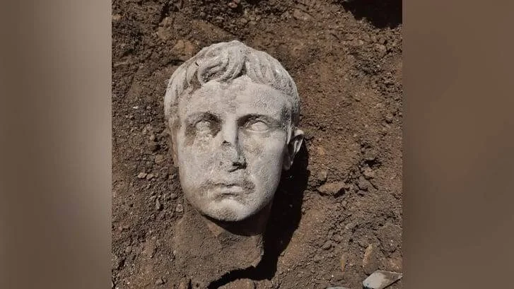 Cabeza de mármol de figura masculina romana recién desenterrada, rodeada de tierra oscura.