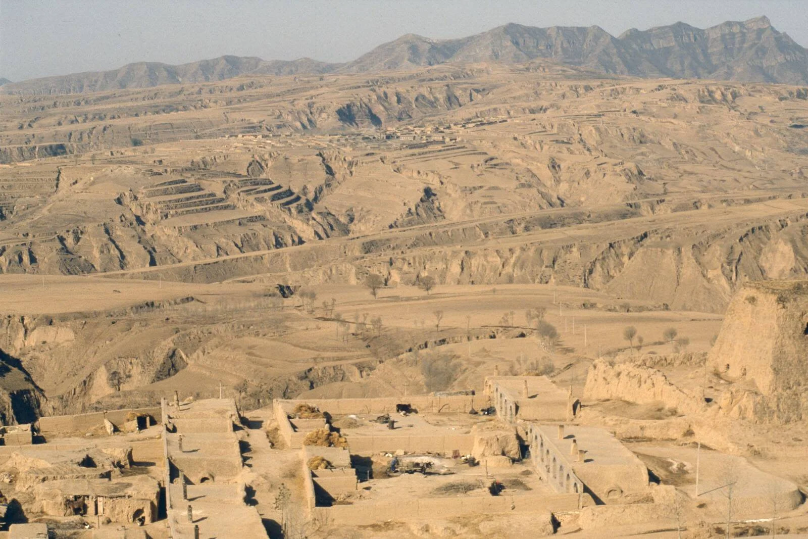 Conjunto de construcciones de adobe en primer plano con un vasto paisaje desértico erosionado y montañas al fondo.