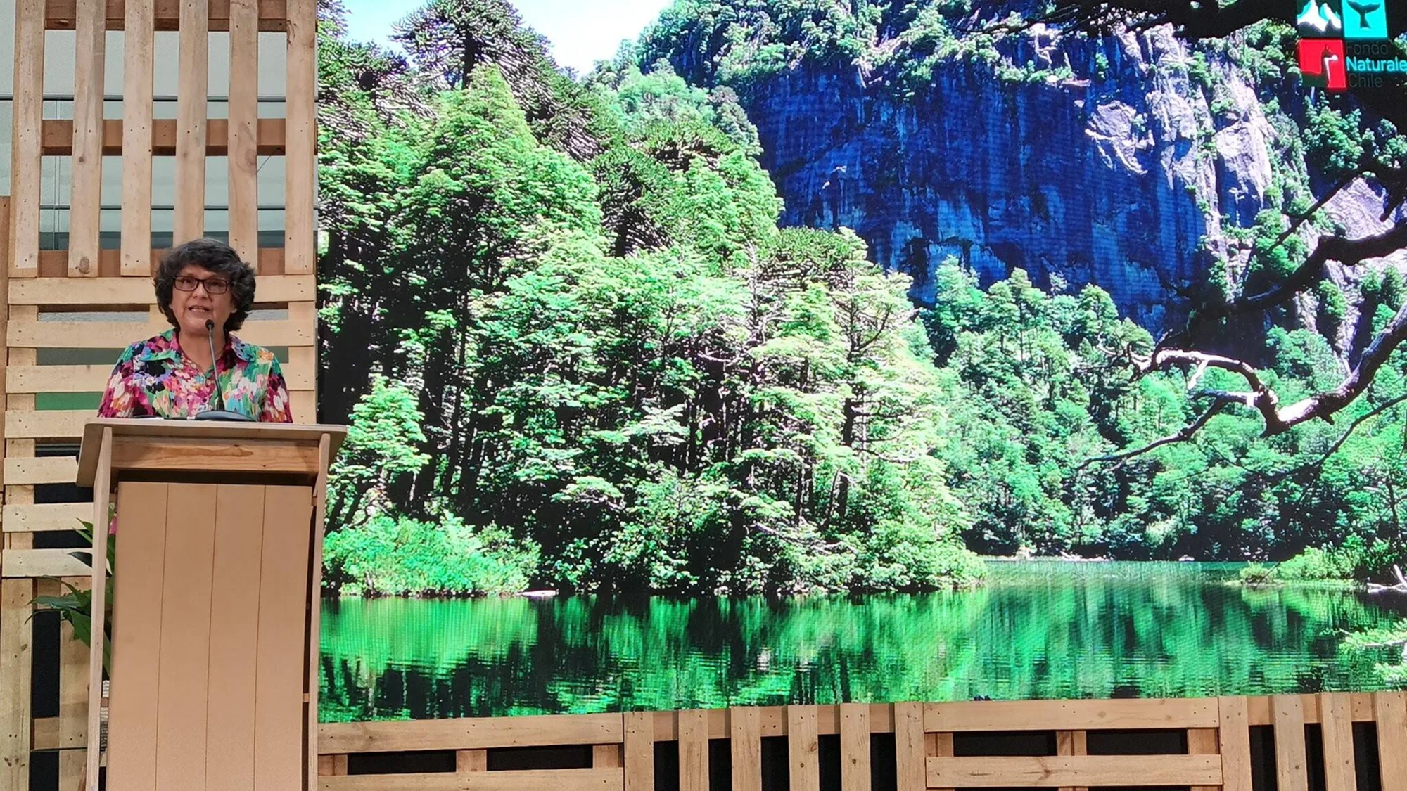 Mujer con blusa floral habla en un podio frente a una gran pantalla que muestra un bosque con lago, en evento de Fondo Natural Chile.