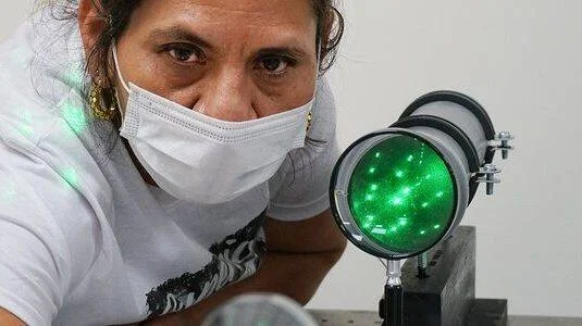 Mujer con barbijo blanco observa de cerca un dispositivo óptico que emite luz láser verde en un laboratorio.