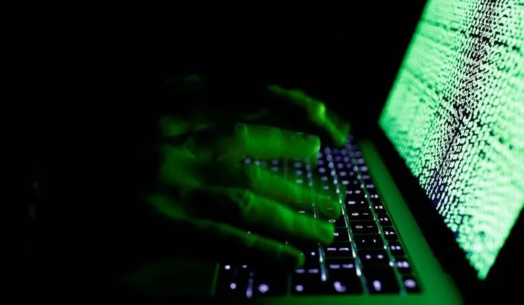 Manos iluminadas con luz verde teclean un portátil que muestra código en pantalla, evocando ciberataque o hackeo.
