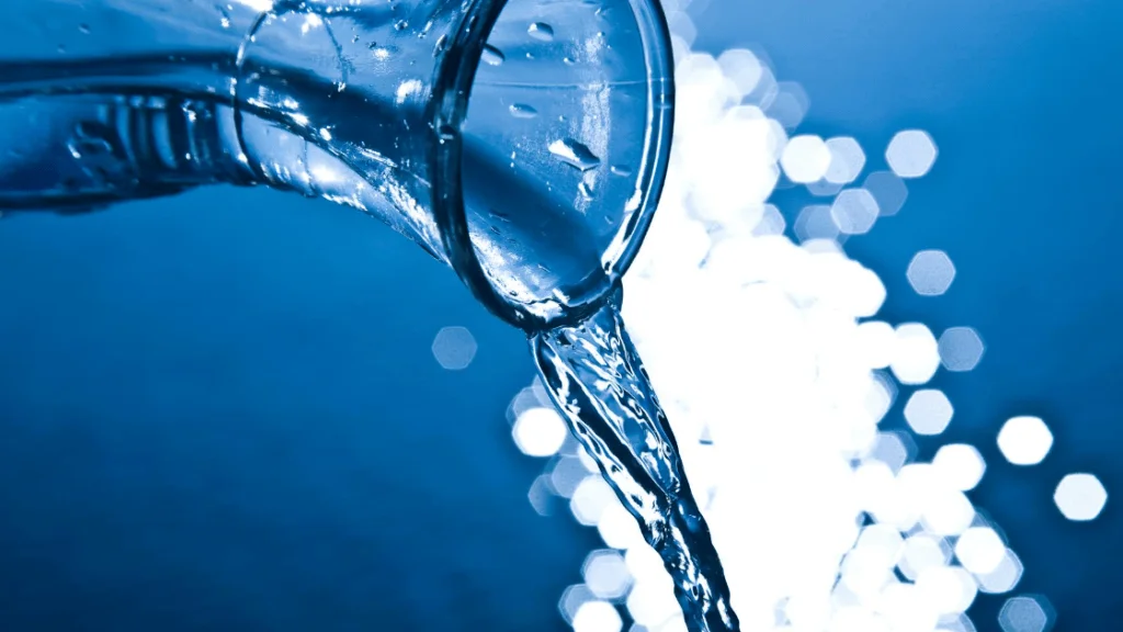 Agua cayendo desde el cuello de una botella de vidrio sobre fondo azul con destellos bokeh.