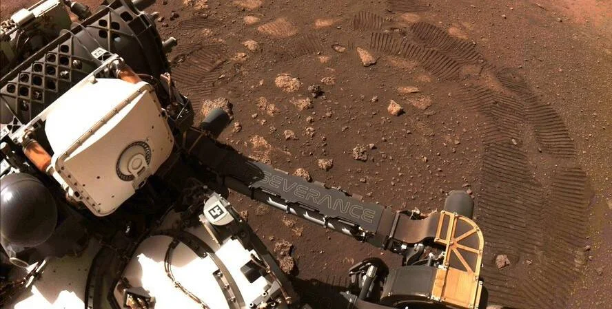 El rover Perseverance sobre la superficie rojiza de Marte, con sus huellas de ruedas visibles en el suelo rocoso.