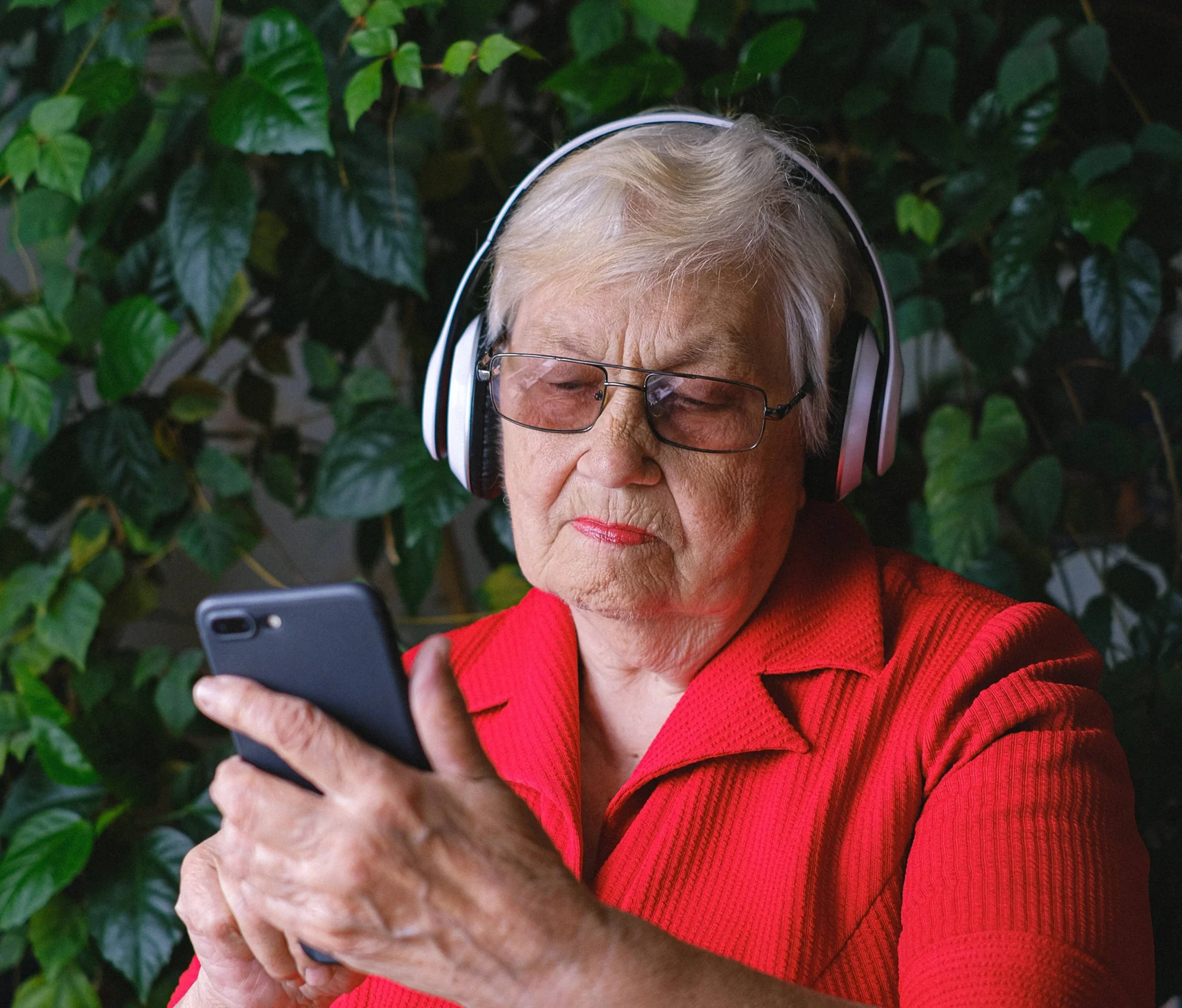 Mujer mayor con auriculares inalámbricos y anteojos revisa su teléfono celular frente a un fondo de plantas verdes.