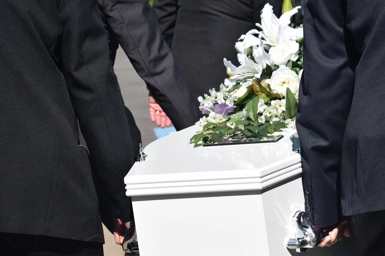 Personas vestidas de negro cargan un ataúd blanco adornado con flores blancas durante un funeral.