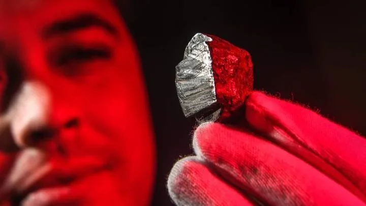 Una persona con guante rojo sostiene un pequeño fragmento de mineral metálico brillante bajo luz roja intensa.
