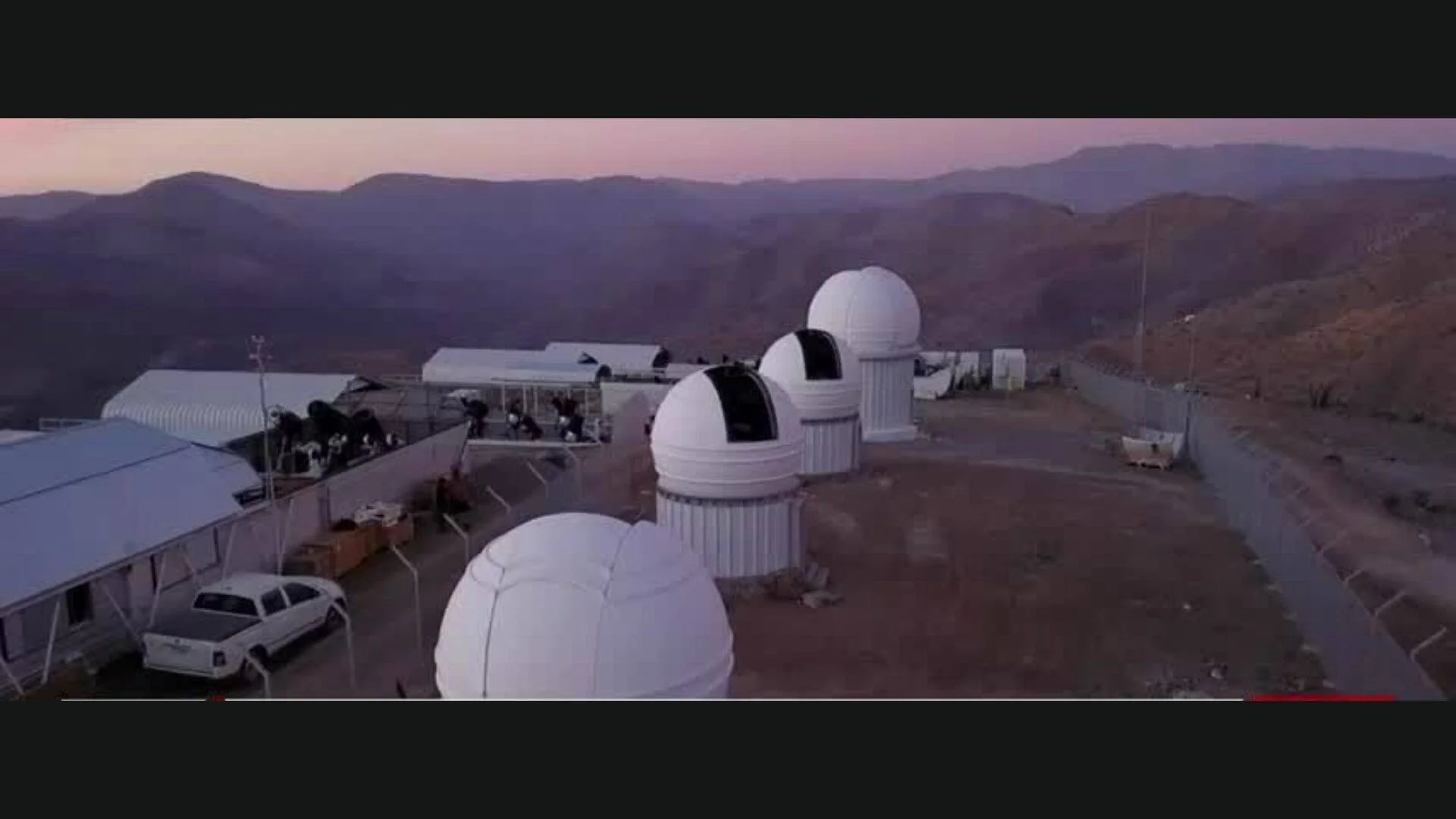 Vista aérea de un observatorio astronómico con múltiples cúpulas blancas ubicado en un terreno árido rodeado de montañas al atardecer.