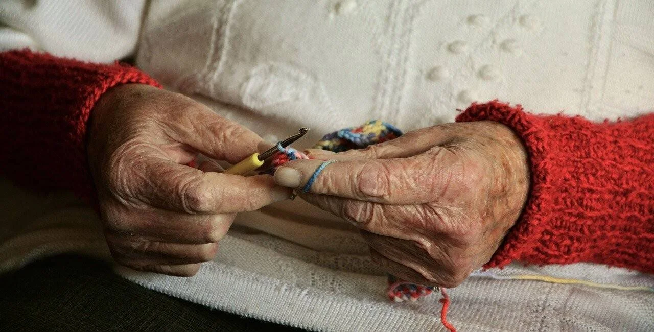 Manos muy arrugadas de una persona mayor con suéter rojo tejiendo a crochet con hilo de colores.