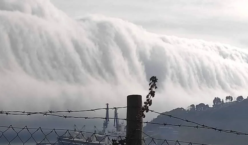 Una densa masa de nubes cae en cascada sobre una montaña hacia un puerto con barcos, vista desde un alambrado de púas.