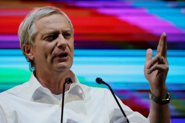 Hombre de cabello canoso habla en un micrófono con la mano levantada, frente a un fondo colorido.