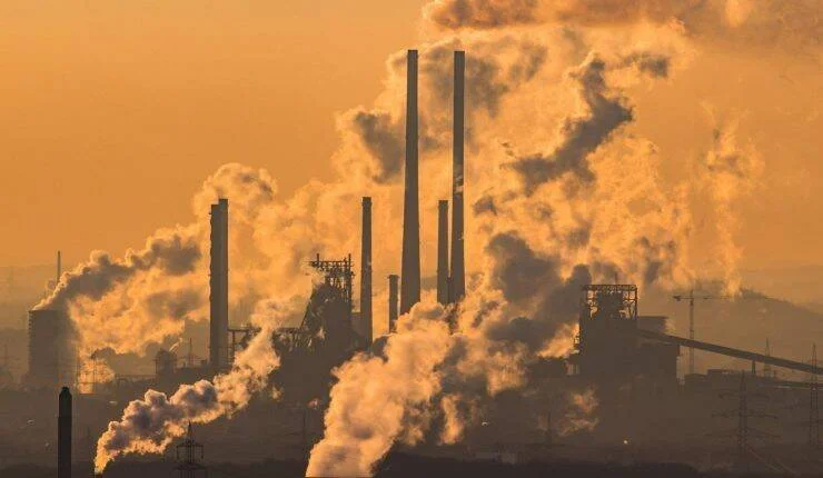 Planta industrial con múltiples chimeneas expulsando grandes columnas de humo bajo un cielo naranja contaminado.