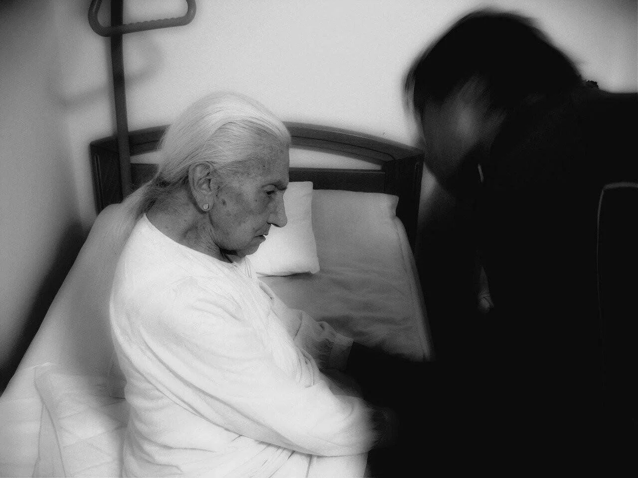 Mujer mayor sentada en una cama de hospital recibe atención de un cuidador, en imagen en blanco y negro de tono intimista.