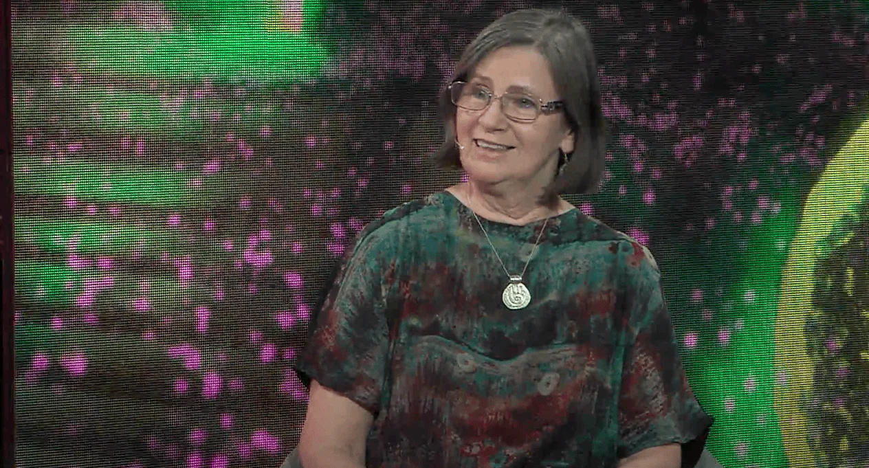 Mujer mayor con anteojos y camiseta verde estampada sonríe frente a un fondo con pantalla LED de colores.