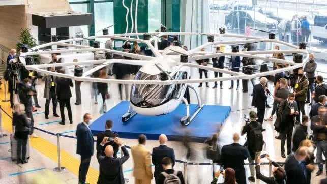 Prototipo de taxi aéreo eléctrico multirrotor exhibido en un salón rodeado de visitantes y periodistas.