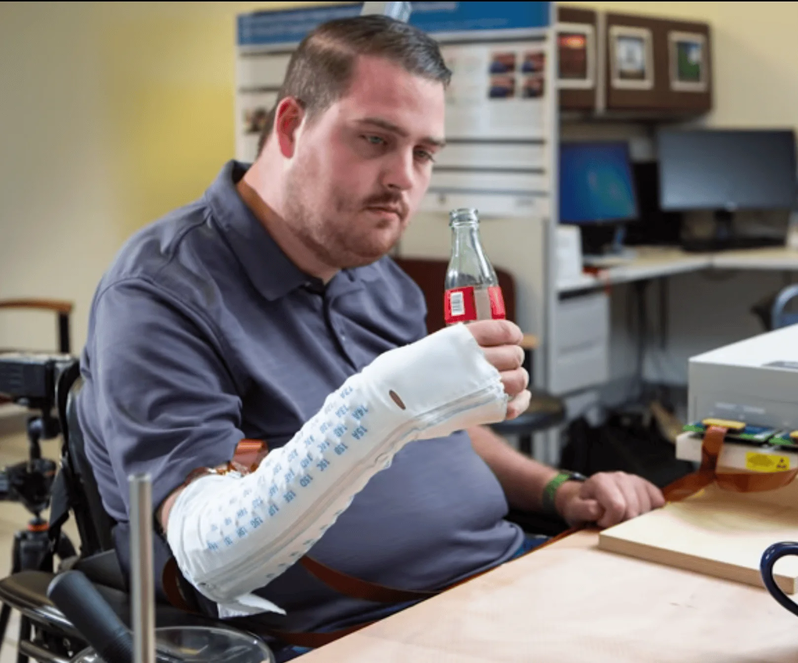 Hombre en silla de ruedas usa un exoesqueleto de brazo para sostener una botella de vidrio en un laboratorio.