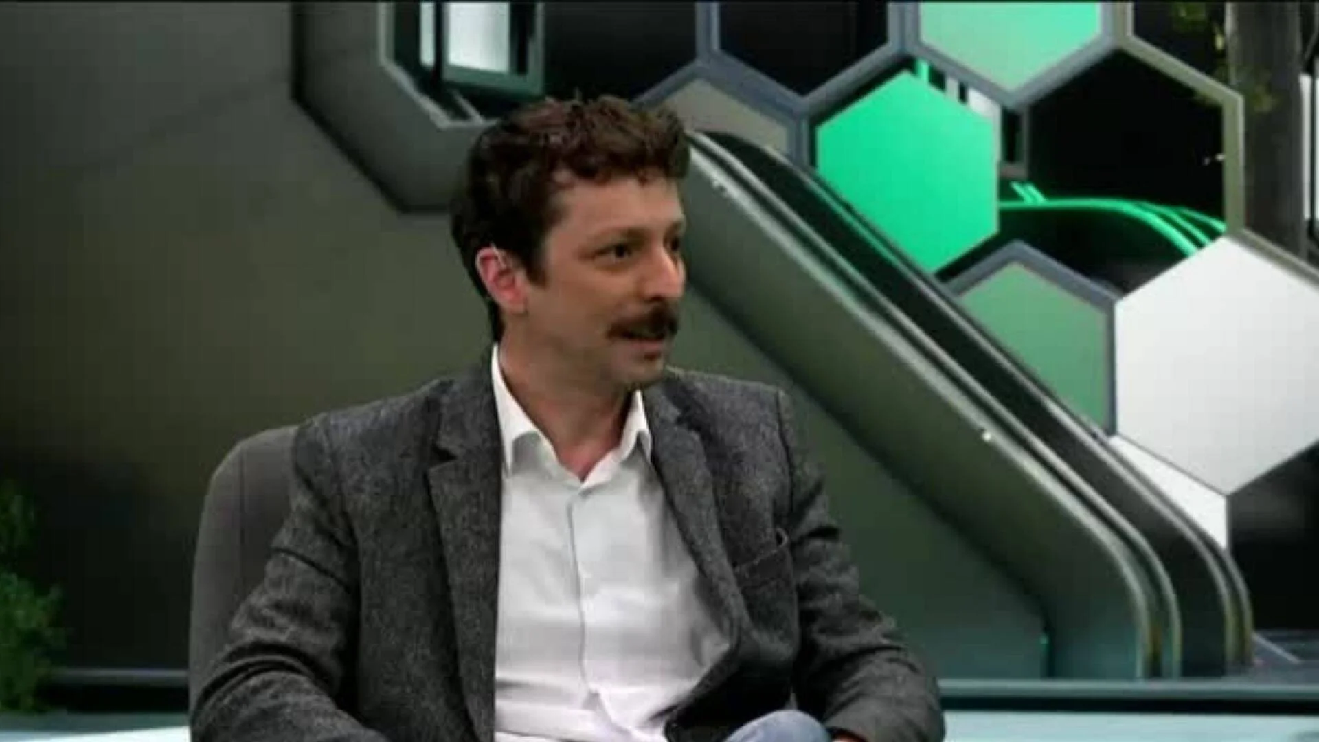 Hombre con saco gris y camisa blanca sentado en un estudio de televisión con decorado de hexágonos verde y gris.