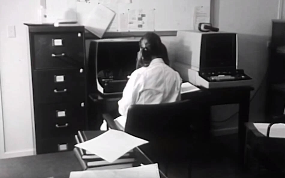 Fotografía en blanco y negro de una mujer de espaldas trabajando frente a una computadora antigua en una oficina con archivadores.