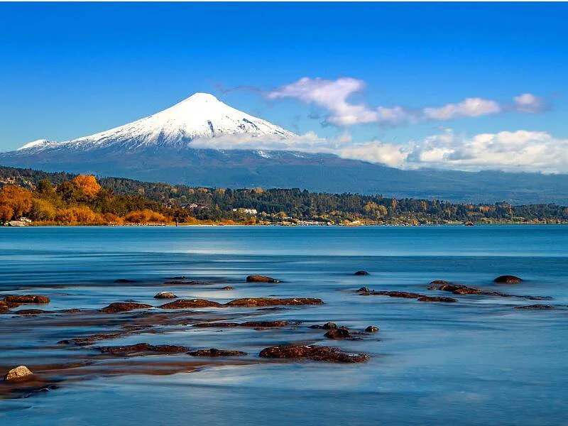 Volcán de cima nevada se eleva sobre un lago de aguas azules con rocas en primer plano y bosque otoñal en la orilla.