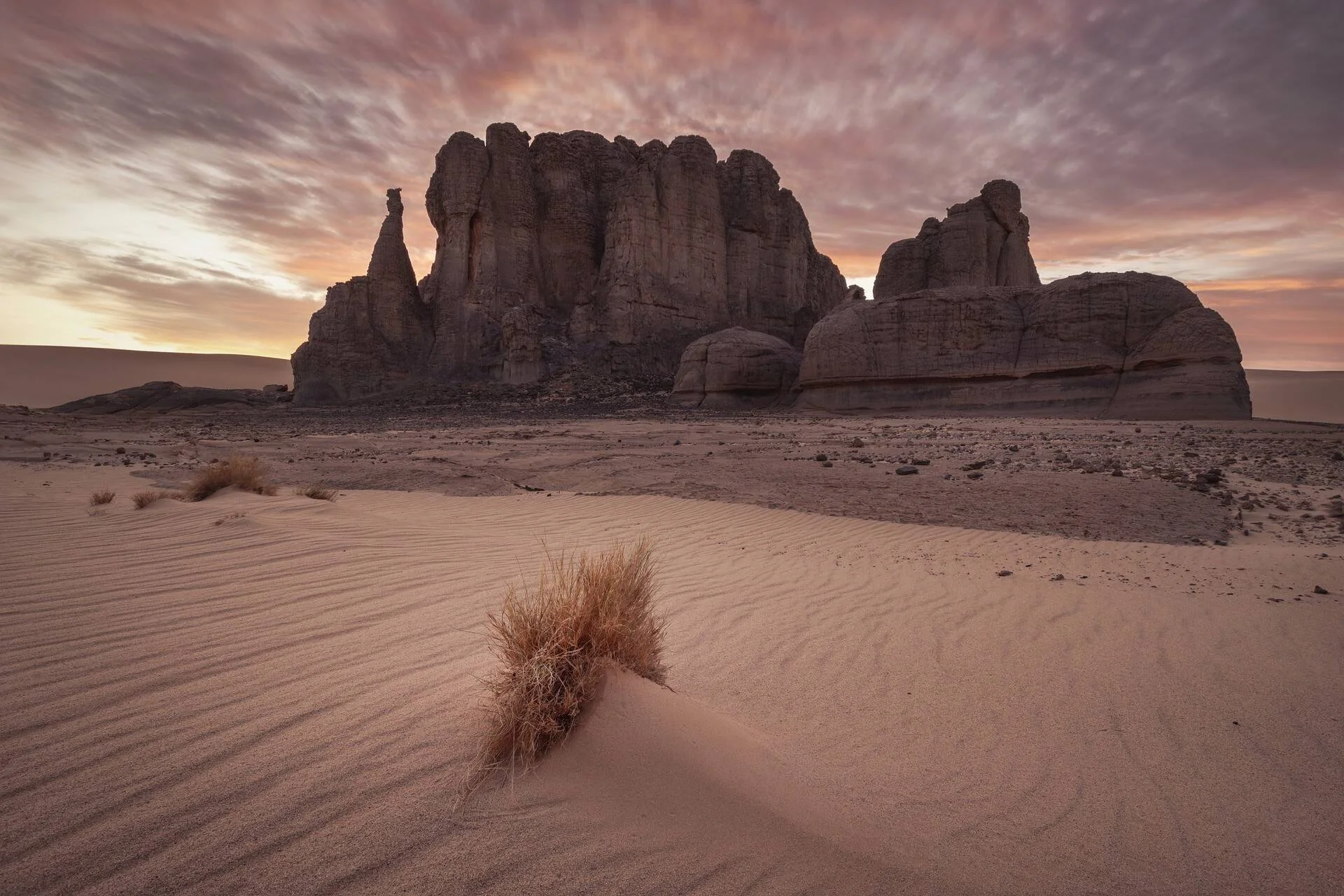 Imponente macizo rocoso erosionado se alza sobre dunas de arena con una mata seca en primer plano bajo un cielo rojizo al atardecer.