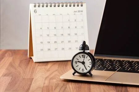 Reloj despertador negro sobre una laptop junto a un calendario de escritorio del mes de junio, sobre una mesa de madera.