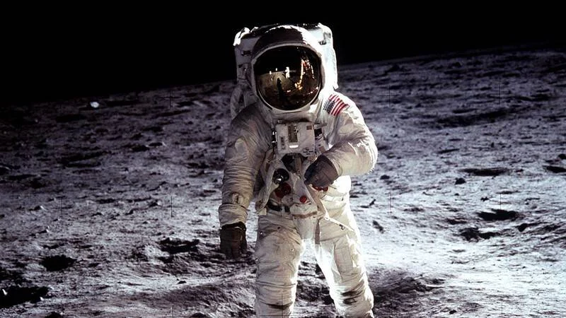 Astronauta con traje espacial blanco y bandera de EE.UU. caminando sobre la superficie lunar.