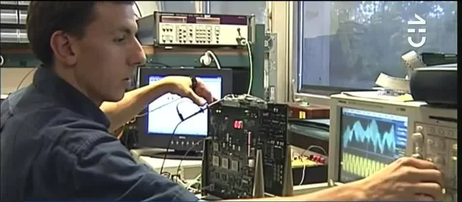 Técnico manipula una placa de circuito impreso rodeado de osciloscopios y equipos de medición electrónica.