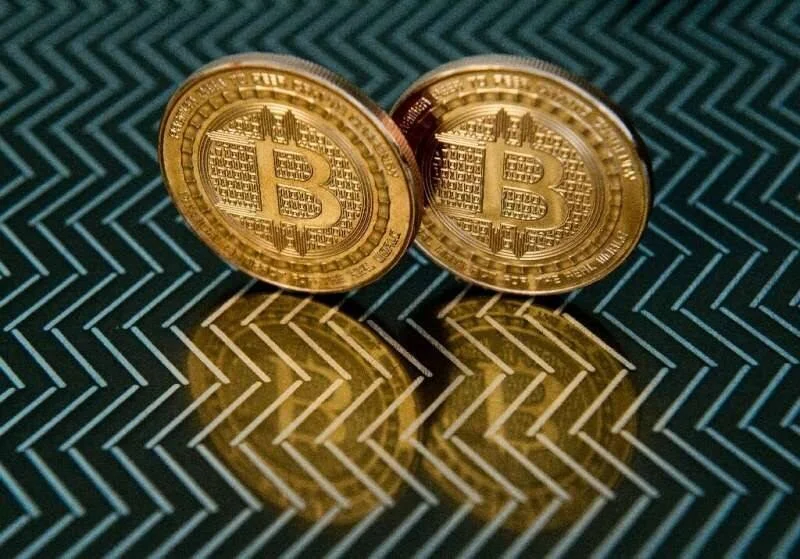 Dos monedas físicas doradas con el símbolo de Bitcoin reflejadas sobre una superficie con patrón geométrico en zigzag.