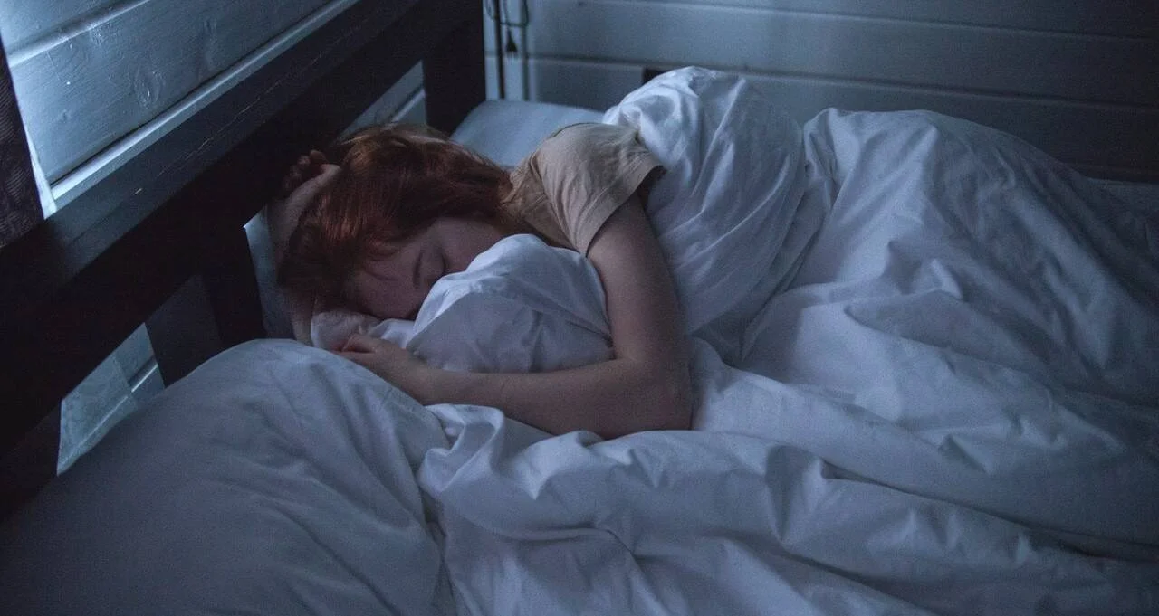 Mujer pelirroja duerme profundamente en una cama con sábanas blancas en un cuarto a oscuras.