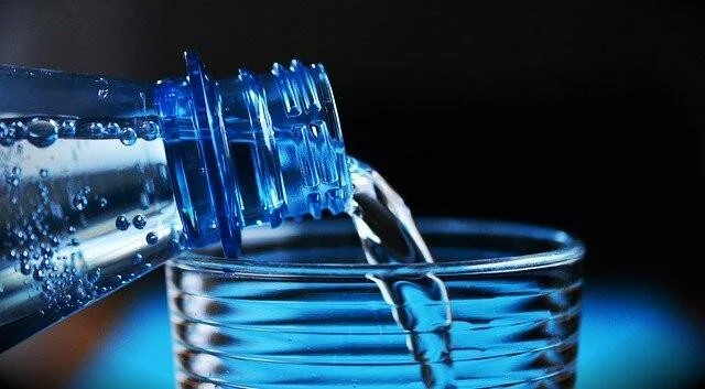 Agua siendo servida desde una botella de plástico azul hacia un vaso de vidrio sobre fondo oscuro.